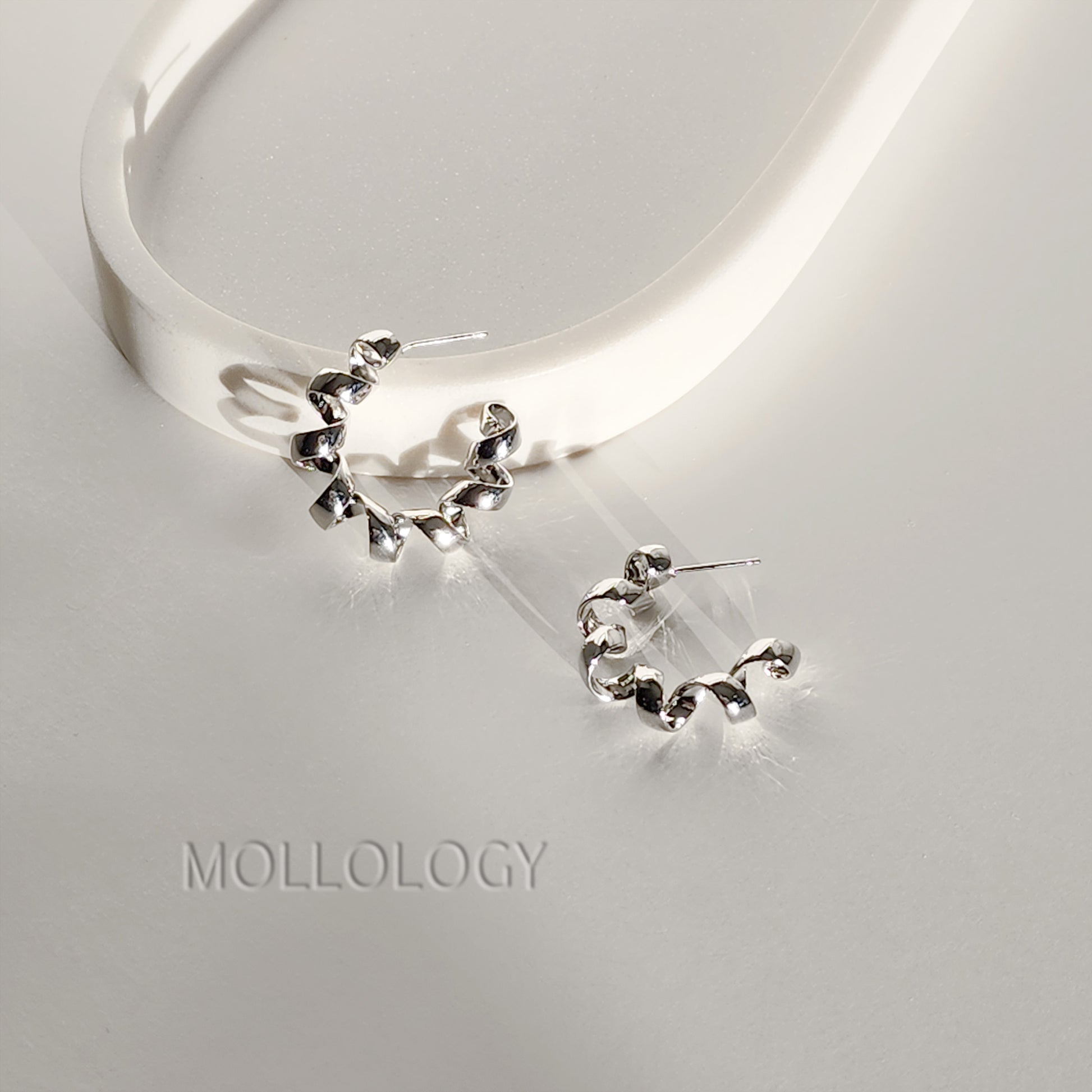 A pair of silver crescent-shaped hoop earrings with a textured, hammered finish, displayed on a light gray surface next to a white curved display stand. The brand name 'MOLLOLOGY' is subtly printed on the surface below.