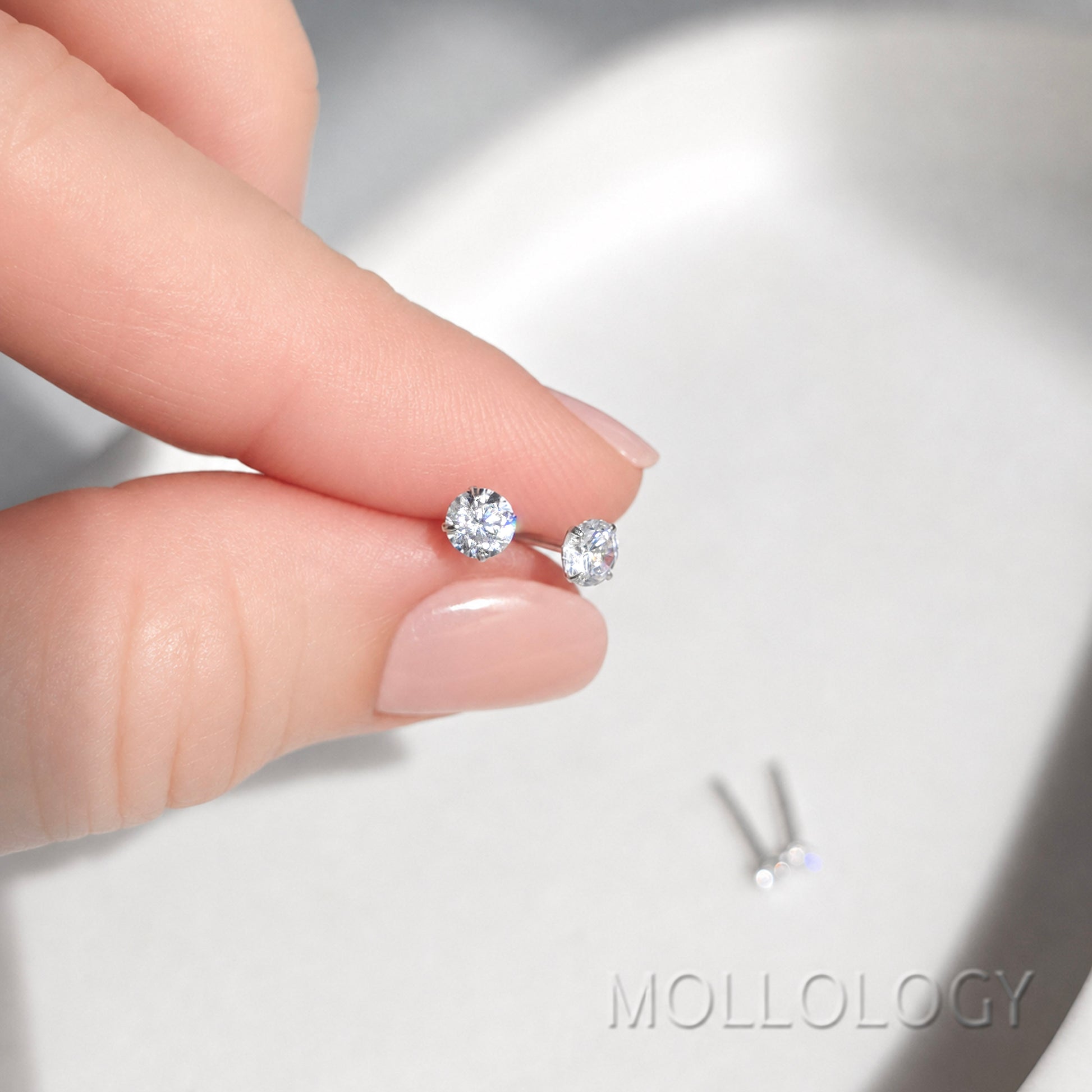 A close-up of a hand holding a pair of small round stud earrings with silver posts and clear gemstones, with the brand name MOLLOLOGY visible in the lower right corner.