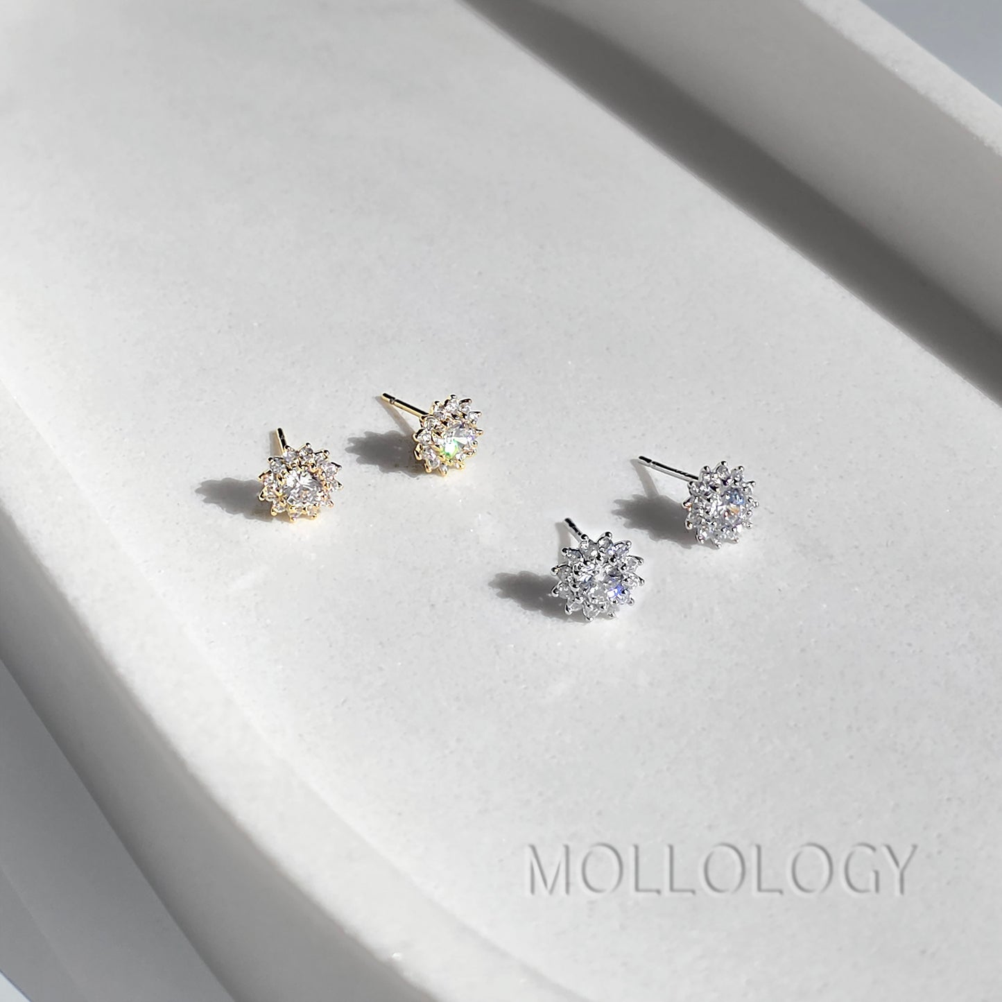 Four snowflake-shaped stud earrings displayed on a white textured surface, featuring both gold and silver metal finishes. The brand name MOLLOLOGY is printed on the surface.