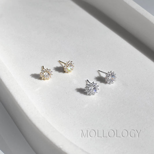 Four snowflake-shaped stud earrings displayed on a white textured surface, featuring both gold and silver metal finishes. The brand name MOLLOLOGY is printed on the surface.