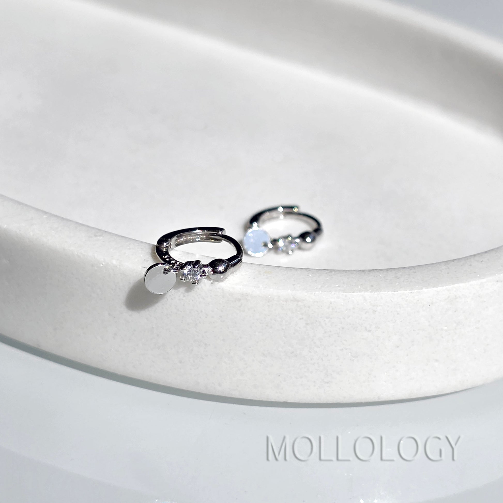 A pair of silver huggie hoop earrings with small clear gemstones and a flat round disc charm, displayed on a white ceramic dish. The brand name MOLLOLOGY is printed on the surface.