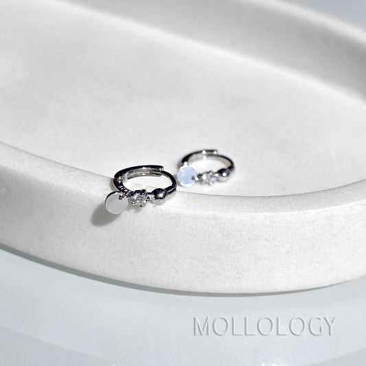 A pair of silver huggie hoop earrings with small clear gemstones and a flat round disc charm, displayed on a white ceramic dish. The brand name MOLLOLOGY is printed on the surface.