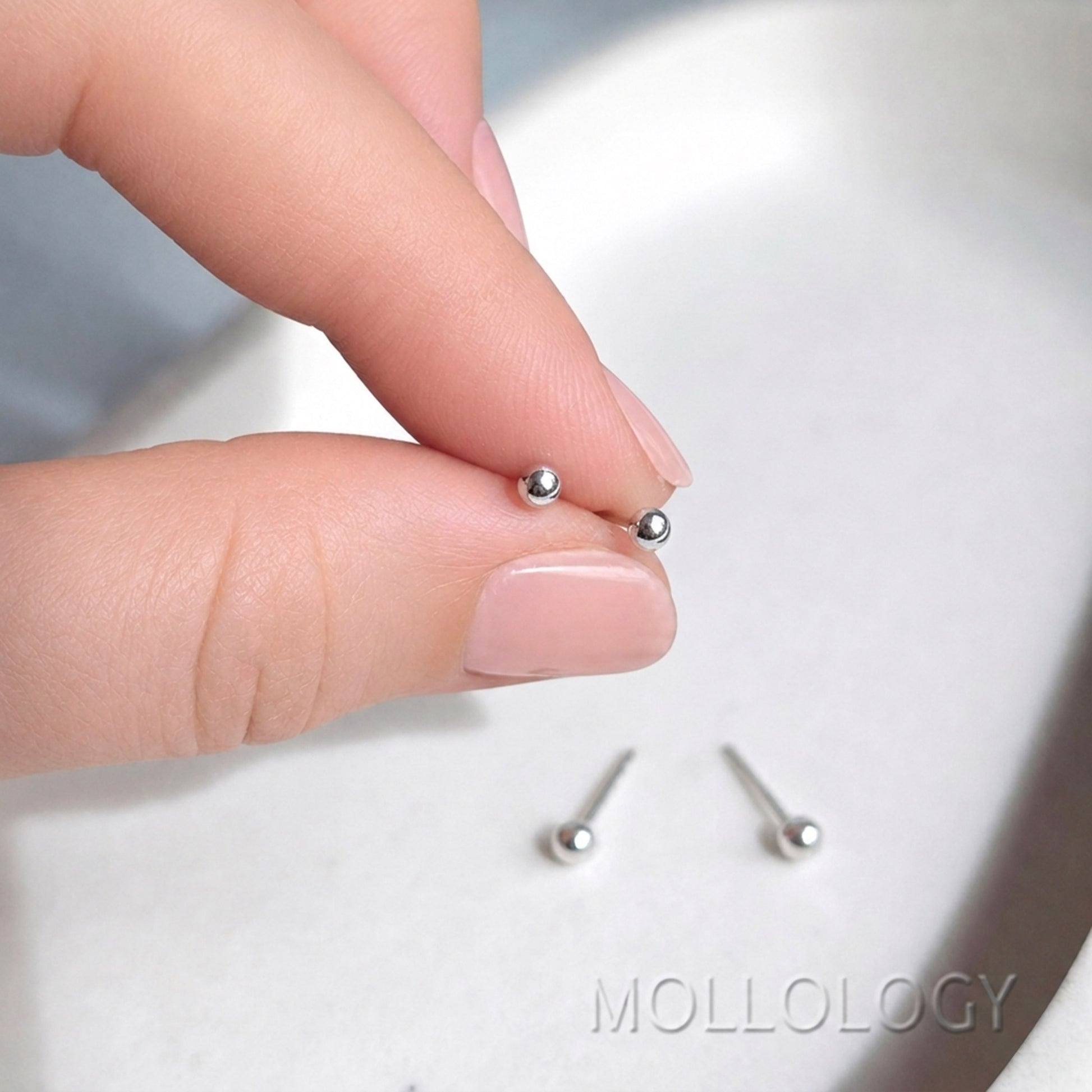 A close-up of a hand holding two tiny silver stud earrings, with two more studs resting on a light surface below. The brand name MOLLOLOGY is visible as a watermark at the bottom right.