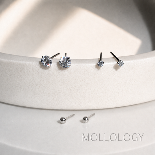 Silver stud earrings displayed on a white curved surface, including floral cluster studs, single round-cut studs, and small ball-back stud earrings. The brand name MOLLOLOGY is printed on the surface.