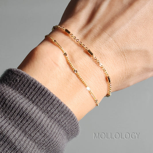 Close-up of a hand wearing a stack of three dainty 14K gold minimalist bracelets, including a satellite bead and bar style chain.
