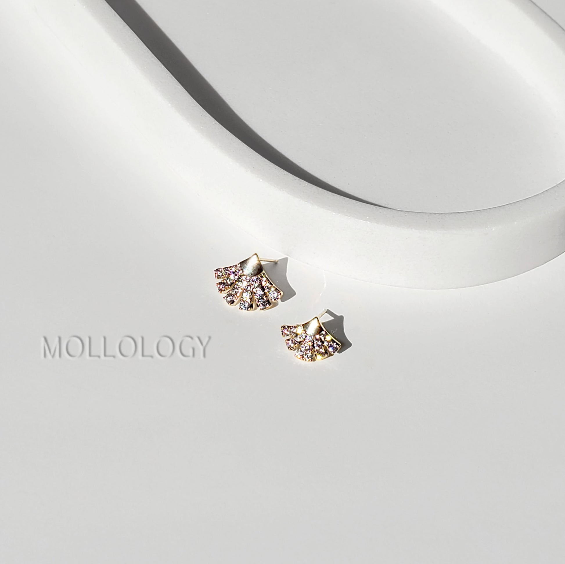 Pair of asymmetrical gold pavé fan-shaped stud earrings with clear sparkling stones on a white sculptural display, with MOLLOLOGY text in the background.