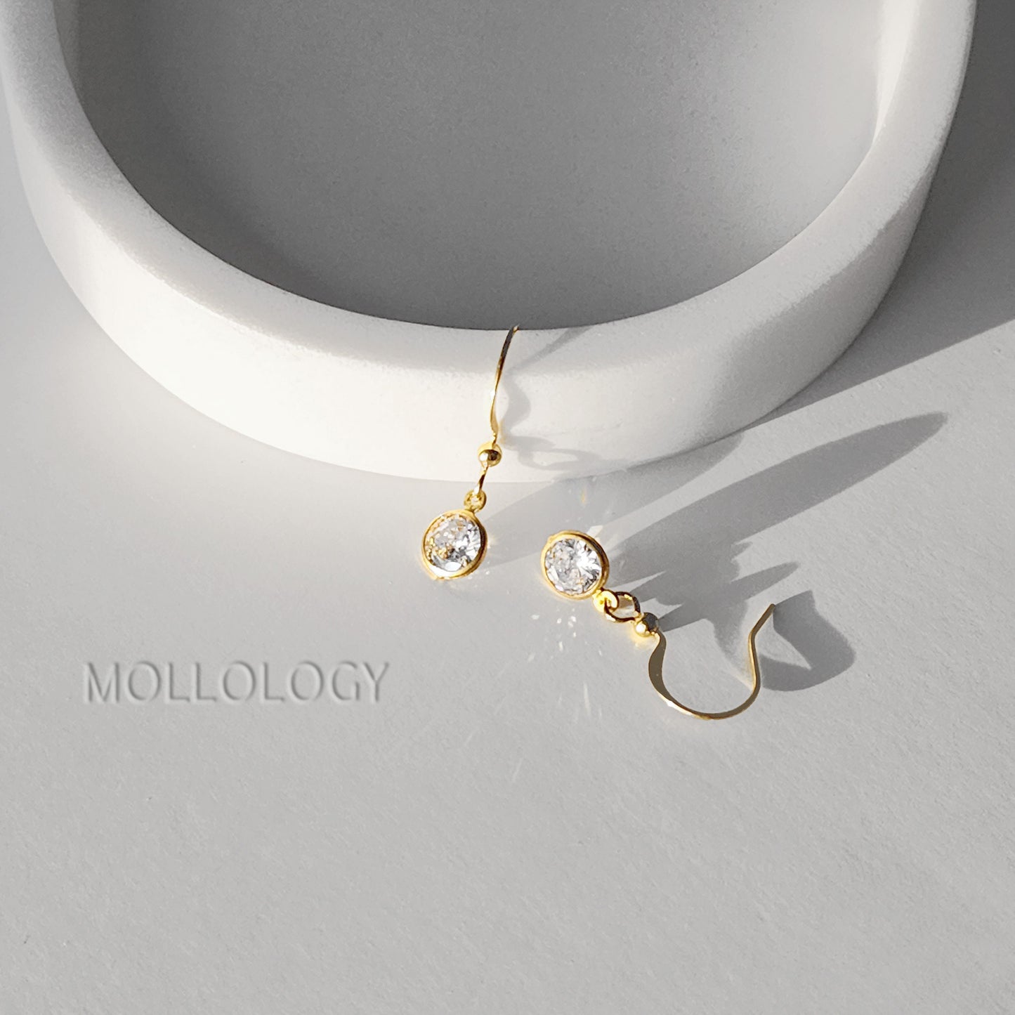 Gold bezel CZ drop earrings on white dish, delicate minimalist dangle earrings casting soft shadows