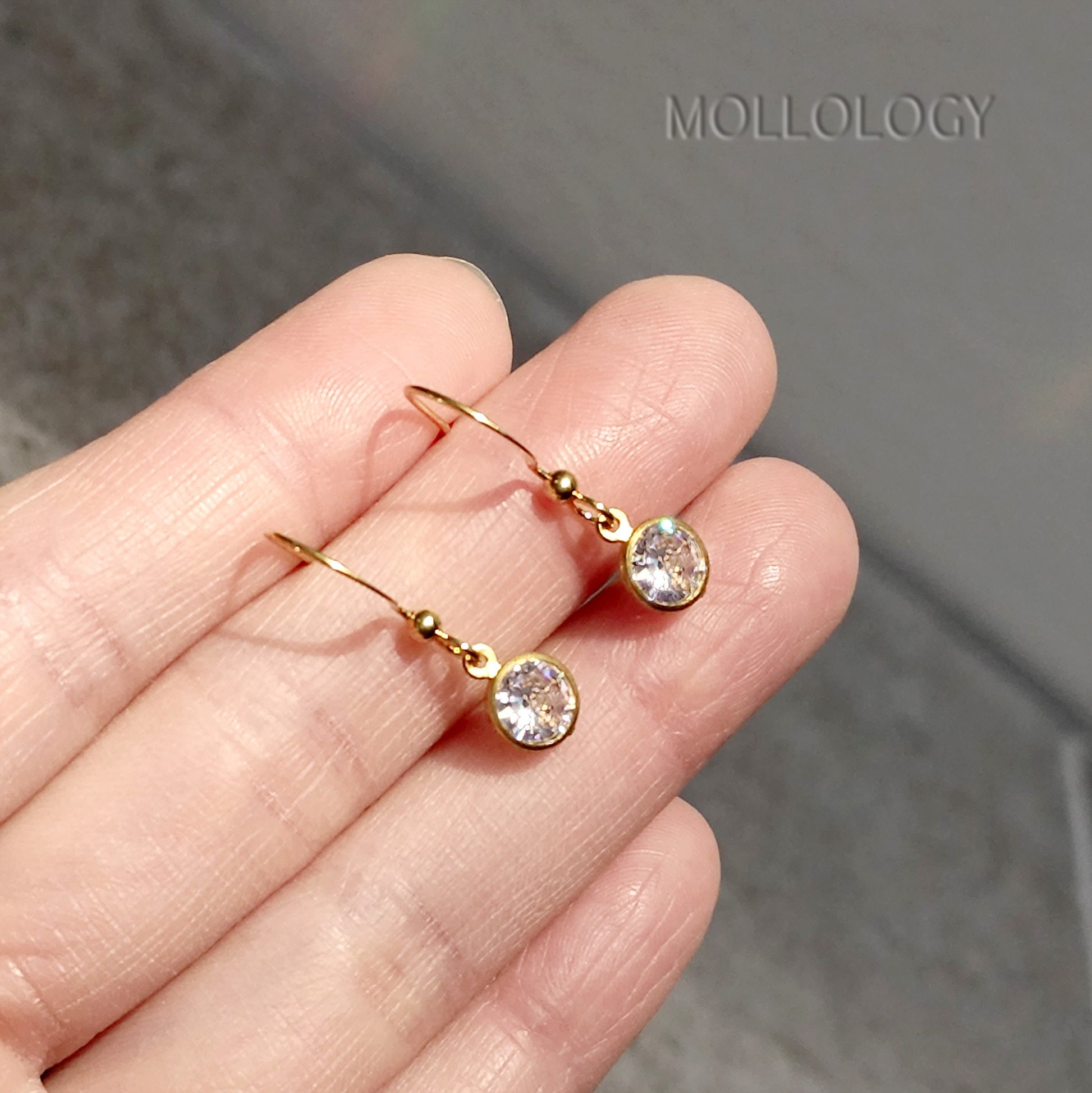 Gold bezel CZ drop earrings resting on fingertips, small minimalist dangle studs for everyday wear