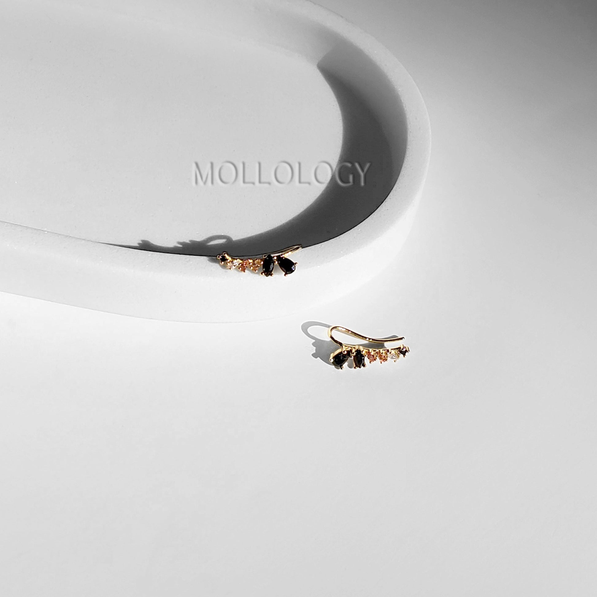 A pair of black onyx and rose quartz ear climbers placed on a white surface with the brand name 'MOLLOLOGY' visible in the background.